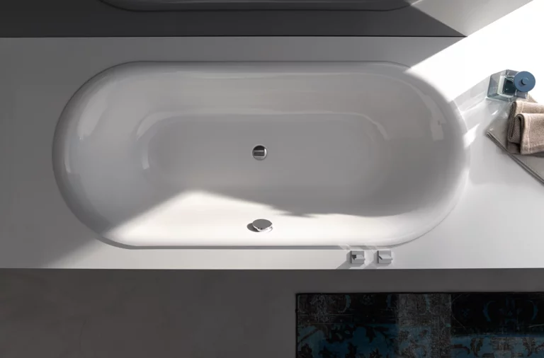 Bañera ovalada de acero esmaltado, integrada en superficie rectangular blanca, con desagüe central.